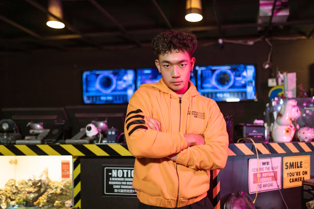 Young male gamer in orange jacket standing confidently in an esports arena with gaming screens in the background.